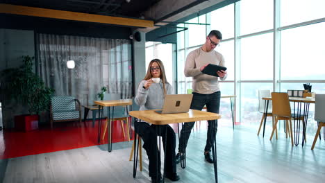 woman sitting at desk in front of laptop drinking coffee. man standing behind is focused on his i-pad.
