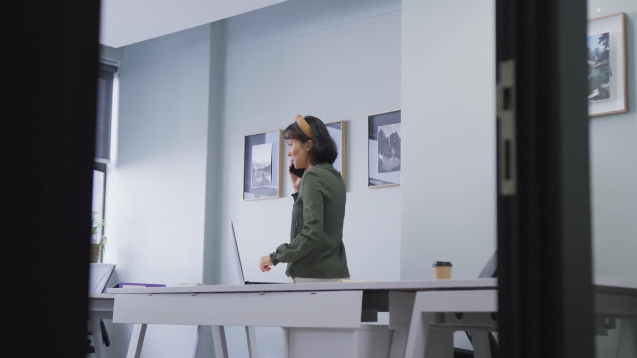 mujer de negocios biracial de pie en la mesa y hablando en el teléfono inteligente sola en la oficina