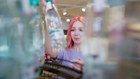 Close-up-a-confident-girl-with-pink-hair-in-a-purple-T-shirt-approaches-a-counter-with-bottles-of-water-and-chooses-the-right-one-for-herself-in-a-supermarket