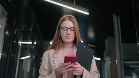 retrato de una mujer de negocios con gafas caminando en el pasillo de la oficina y respondiendo en un chat de trabajo de teléfono móvil