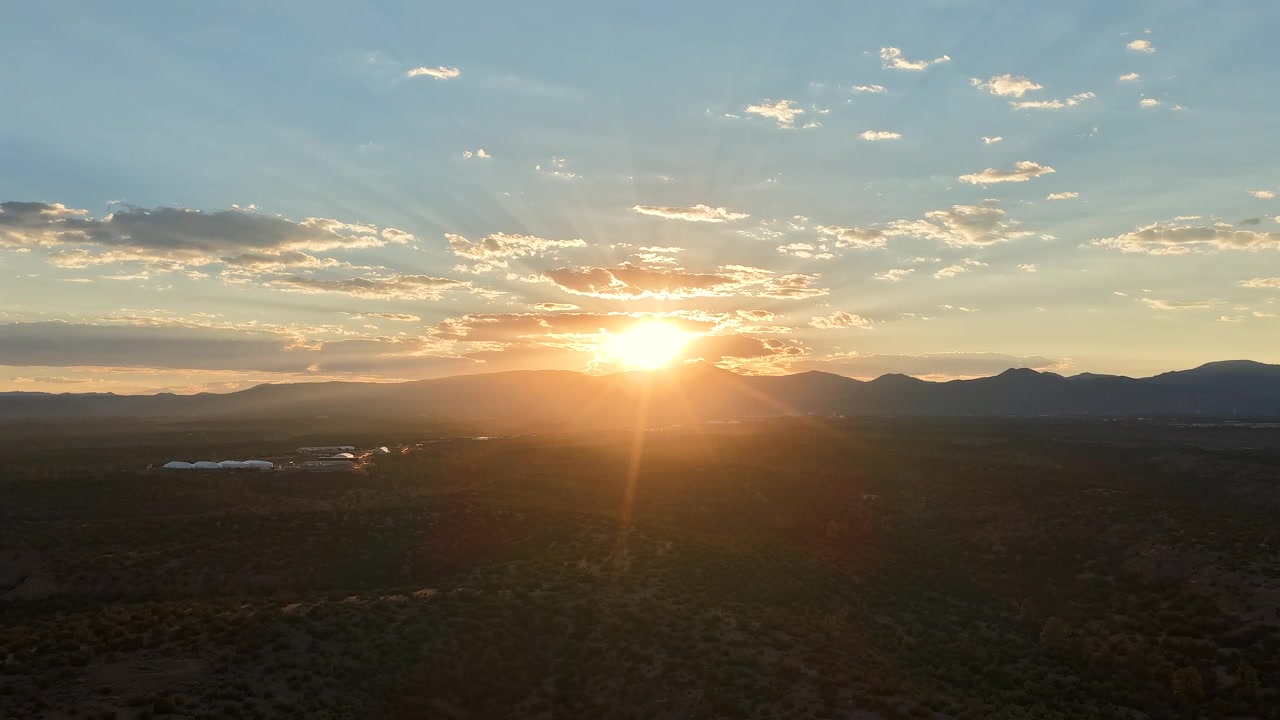 4K drone hyperlapse capturing a golden sunset over the dark mesas of White Rock, New Mexico. Radiant sunbeams burst through the clouds above the distant silhouette of the Jemez Mountains.