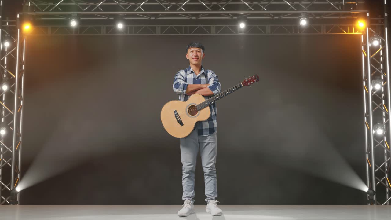 cuerpo lleno de adolescente asiático con guitarra cruzando los brazos y sonriendo en el escenario