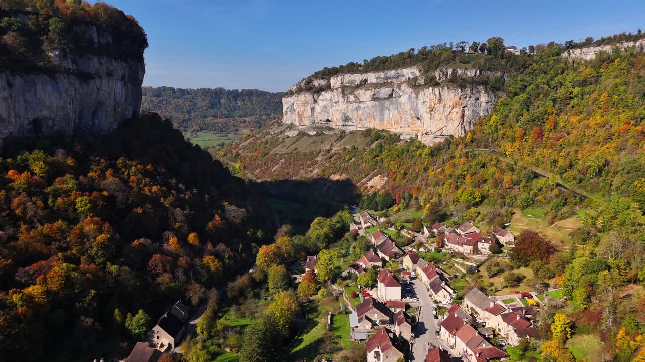 Aerial view of Baume les Messieurs, France, with limestone cliffs, a winding valley and a quiet village surrounded by autumn forests and rolling hills under blue skies in a peaceful rural landscape