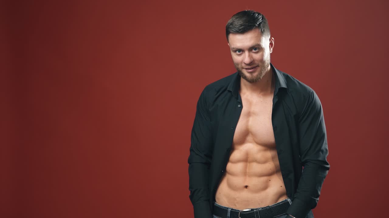 Sexy man in black shirt. Handsome male unbuttons his shirt and shows his naked torso on dark red studio background. Bearded man with sporty body indoors.