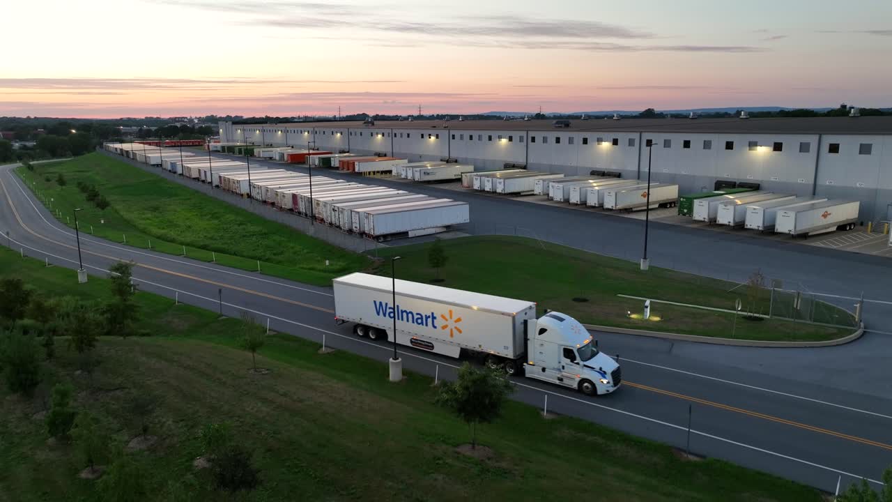 Aerial view of Walmart Truck on rural road in front of warehouse with row of parking semi-trailers. Sunset time in american suburbia. Descending wide shot.