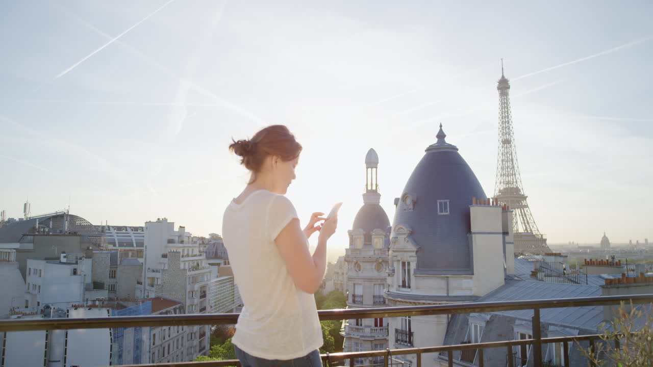 mujer feliz usando un teléfono inteligente enviando mensajes de texto en un balcón en parís francia disfrutando de la vista de la torre eiffel compartiendo experiencias de vacaciones navegando por las redes sociales hermosa puesta de sol