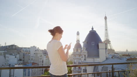 Glückliche-Frau,-Die-Ihr-Smartphone-Nutzt-Und-Auf-Dem-Balkon-In-Paris,-Frankreich,-SMS-Schreibt-Und-Den-Blick-Auf-Den-Eiffelturm-Genießt-Und-Ihr-Urlaubserlebnis-Beim-Surfen-In-Den-Sozialen-Medien-Teilt.-Wunderschöner-Sonnenuntergang