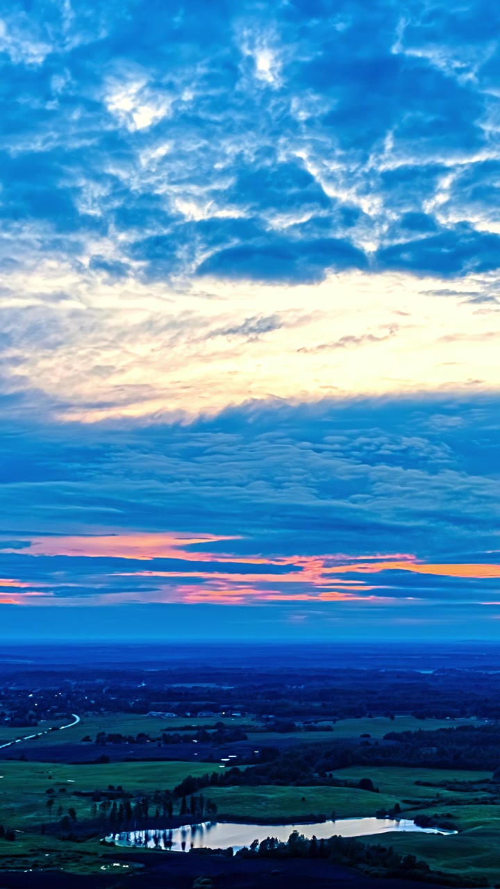 Filmato timelapse verticale mostra un tramonto all'ora d'oro con nuvole stratificate sopra un tranquillo stagno di campagna e luci distanti.