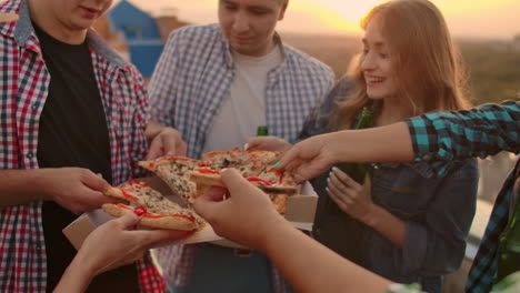 a company of six young russian people in plaid shirts parses pieces of hot pizza with red paper and eats together. this is a rooftop party with a beer.