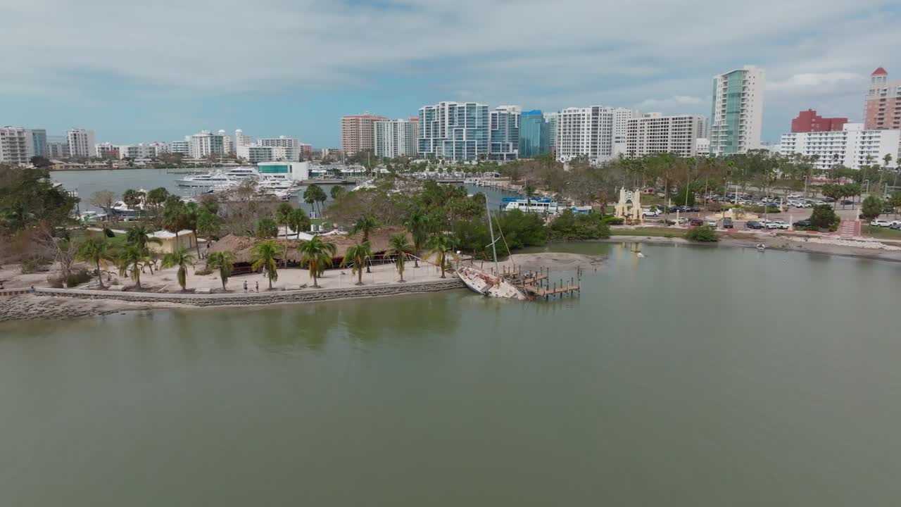 Aerial view of wrecked sailboat grounded at Bayfront Park, Sarasota. Bow submerged, crashed into dock with Marina Jack and downtown Sarasota skyline in the background.