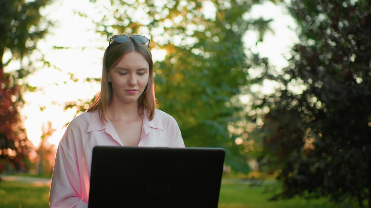 dama de top rosa sentada al aire libre trabajando con una computadora portátil, la luz del sol creando un efecto radiante dorado a su alrededor, con árboles y luz solar filtrándose a través de ellos en el fondo