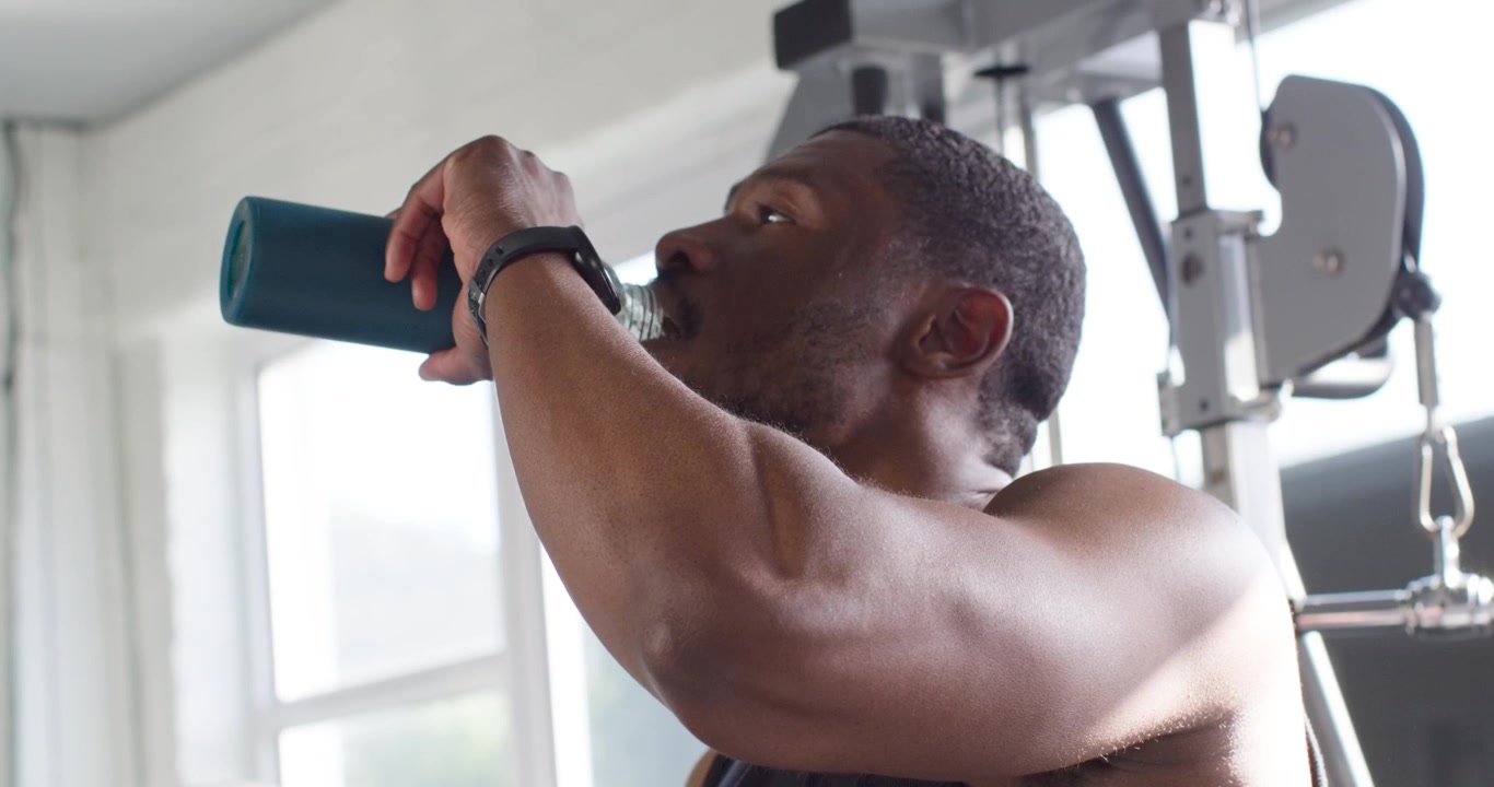 Mid 30s African man ending set opening teal bottle showing yellow tab drinking recovering at gym