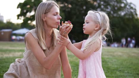 Portrait-Of-Mother-And-A-Small-Daughter,-Spends-Time-Together-In-A-City-Park-On-A-Picnic-They-Bite-The-Little-Cake-From-Sides-Together