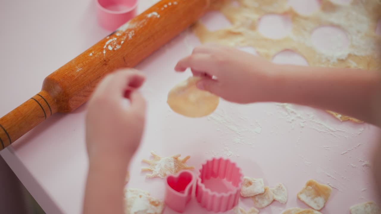 Child releases rolling pin and reaches to pick up small round piece of dough on pastel pink table surrounded by flour, baking cutters, and flattened dough during fun kitchen activity