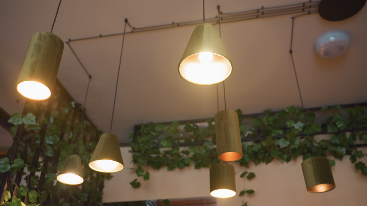 Close up of glowing pendant lights with green fabric shades hanging from ceiling in cozy interior adorned with leafy green plant decor, creating a warm inviting atmosphere