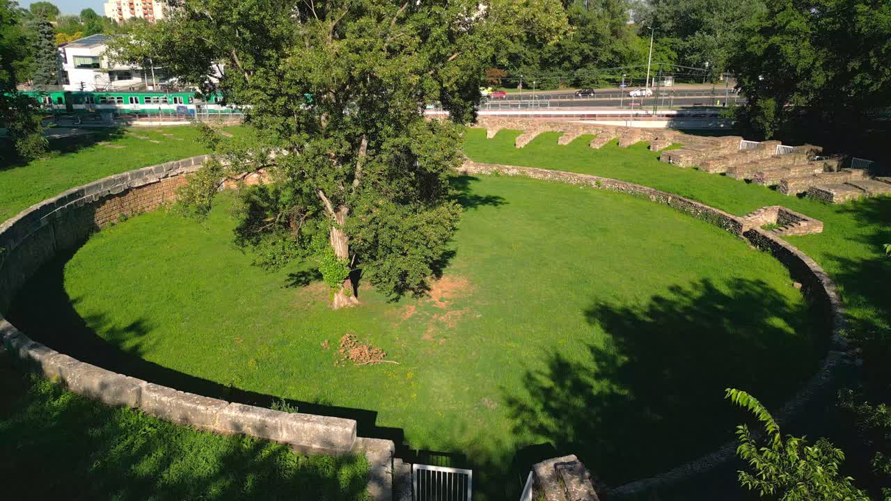 árbol en el medio del anfiteatro civil de aquincum con tren y tráfico en el fondo en budapest, hungría