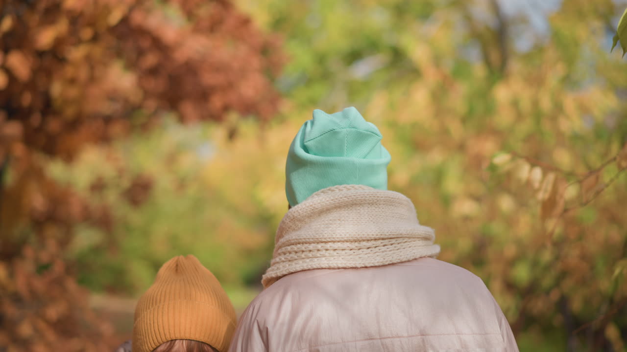 kid and adult bundled in winter jackets and hats walking together on leafy path lined with vibrant autumn trees, both glancing back with curiosity as soft sunlight filters through colorful foliage