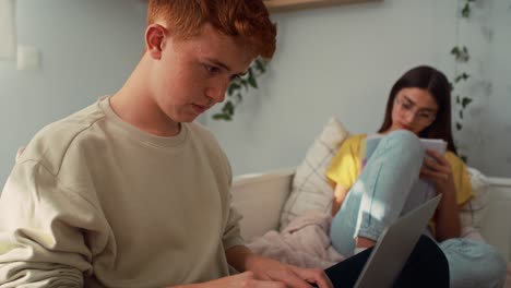 Two-caucasian-teenagers-sitting-on-bed-and-learning-from-books-and-laptop