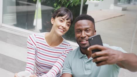 Happy-diverse-couple-wearing-blouse-and-shirt-and-taking-selfie-with-smartphone-in-garden