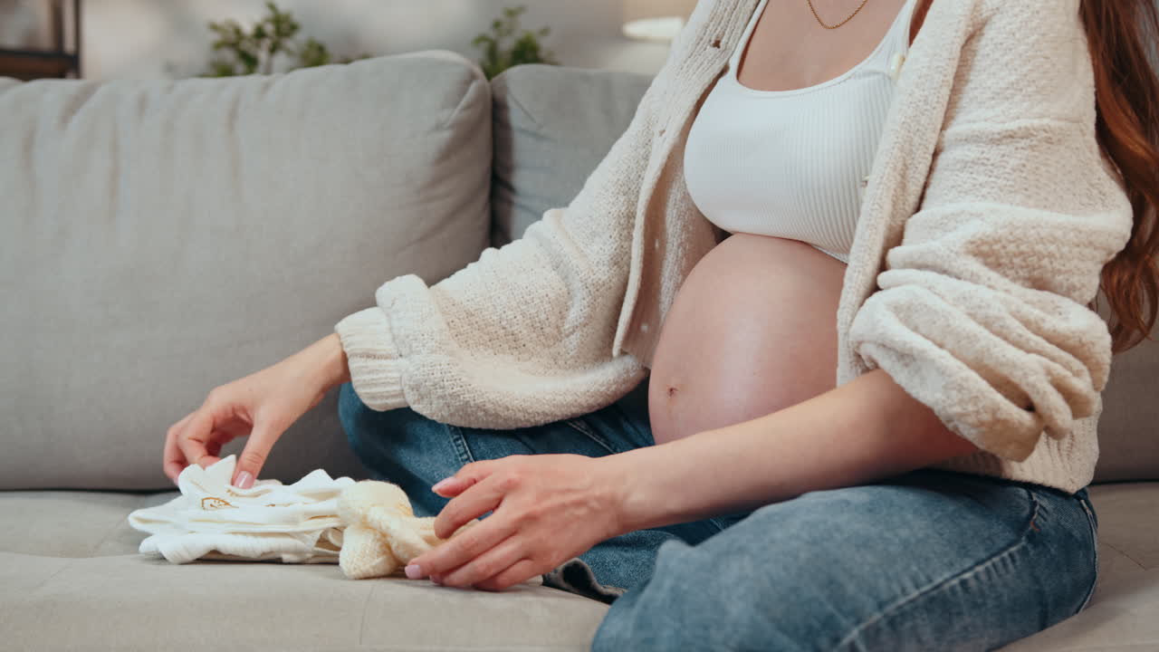 Pregnant woman sitting on couch folding tiny baby clothes and preparing for newborn arrival with