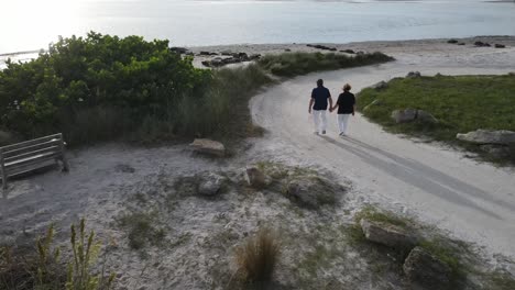 Hermosa-Vista-Aérea-De-Una-Pareja-De-Ancianos-Tomados-De-La-Mano-Mientras-Caminan-Hacia-Una-Playa-Aislada-Al-Atardecer