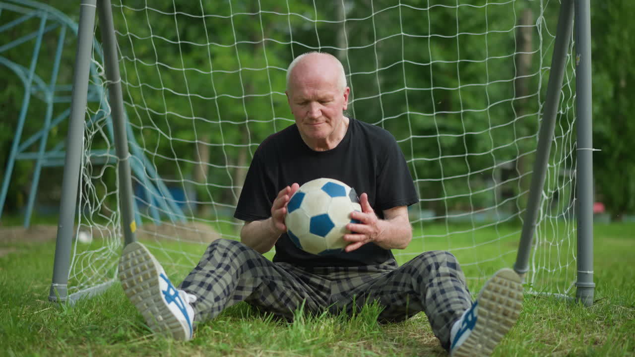 An elderly man sits with open legs near a goal post, lifts a soccer ball that comes towards him, observes it thoughtfully, and then throws the ball back