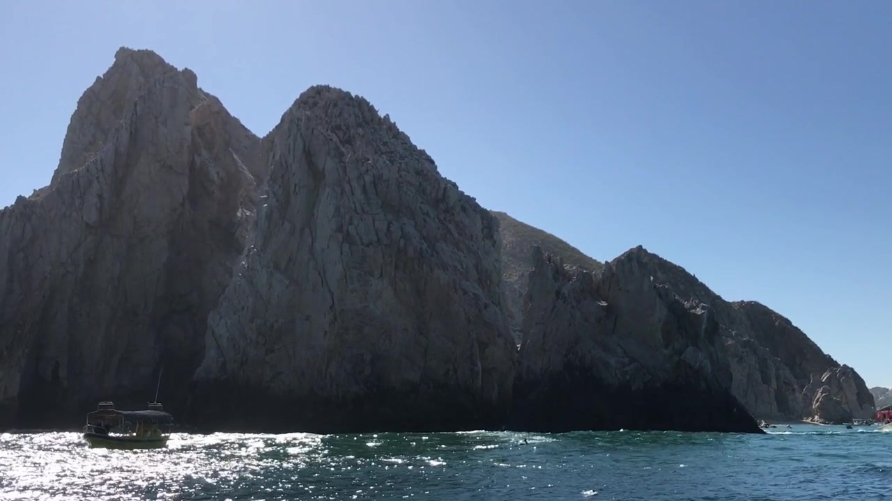 Scogliere costiere rocciose che si ergono sull'oceano blu vicino a Cabo San Lucas, Messico. Piccole imbarcazioni passano lungo la costa frastagliata sotto un cielo azzurro e limpido in pieno giorno