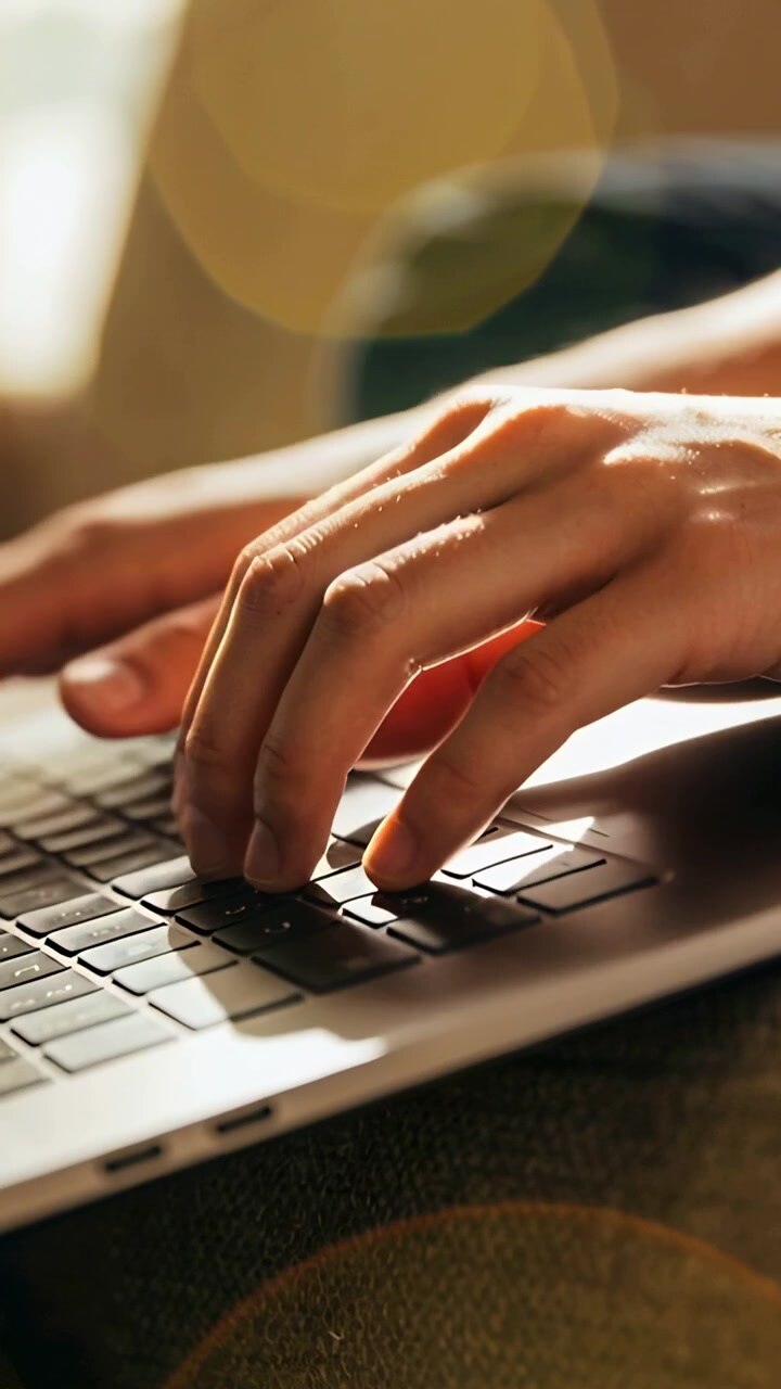 Vertical video: Typing hands from rest pressing laptop keys on lap by window with bare arms, bokeh