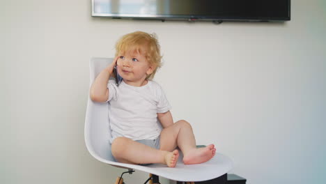 little son in t-shirt and bloomers sits on chair with phone