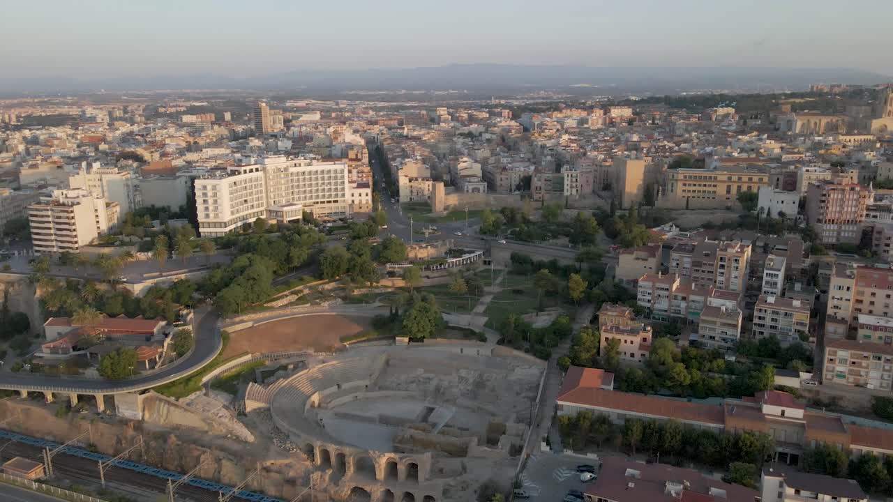 A cinematic slow drone approach to the morning cityscape of Tarragona, Spain, featuring the iconic Roman coliseum and the city's grand cathedral