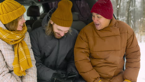 three happy friends talking and laughing together while sitting in car boot on a snowy winter day