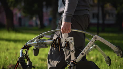 person wearing dark gray pants and matching sweatshirt grips curved metallic bar, possibly part of walking device or stilts, outdoors on blurred green grass during daytime; low camera angle captures