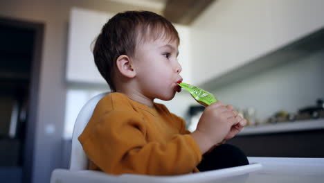little boy having a snack from the doy pack. kid eating puree fruit at home. low angle view.