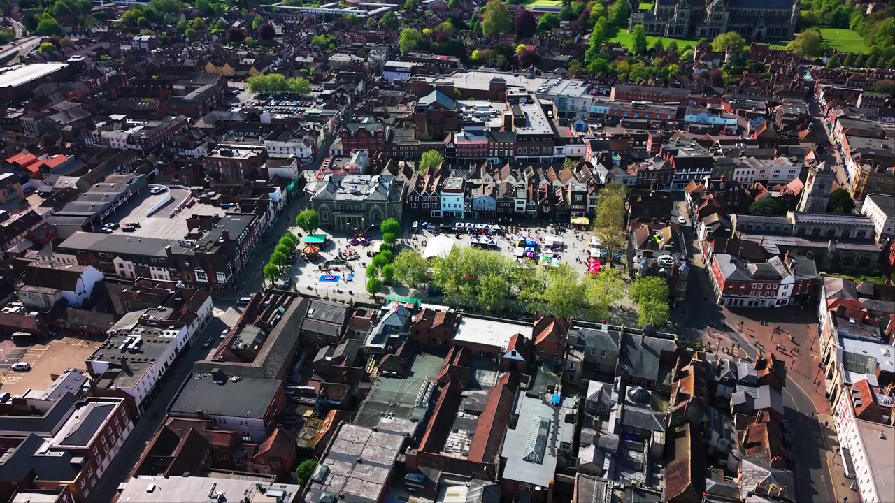 Riprese aeree con drone che si allontanano da Salisbury Market Square durante un evento del Giorno di San Giorgio, rivelando il centro città più ampio, la Guildhall, le bancarelle del mercato, le giostre, la folla, i tetti e le strade.