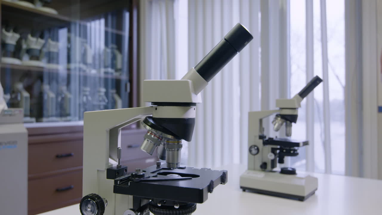 Two microscopes are on a classroom or laboratory counter. One is in the foreground, and the other is in the background.
