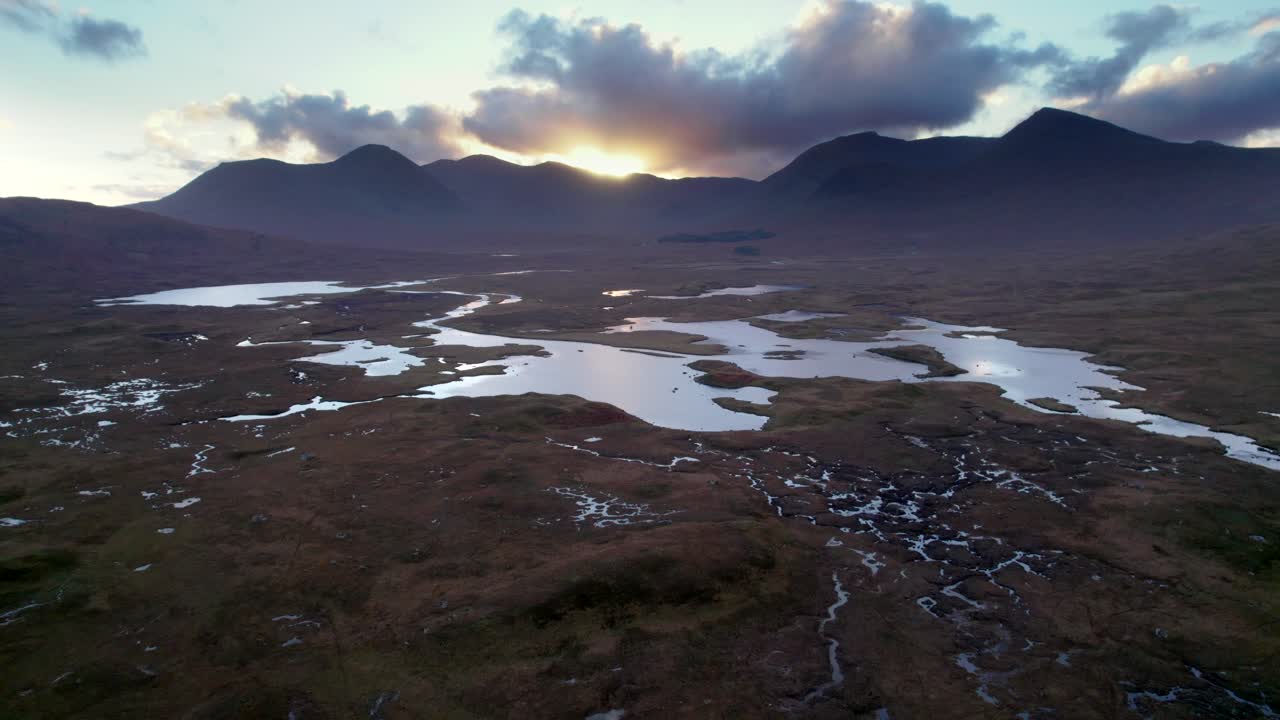 A drone reverses high above a peatland landscape of peat bogs amongst a mosaic of fresh water lochs looking towards dark mountains on the horizon at sunset