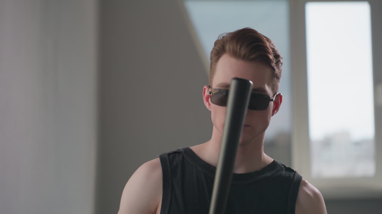 Young guard in black tank and dark sunglasses examines sleek security baton by loft window, muscles flexed in soft daylight, conveying readiness and modern protective style