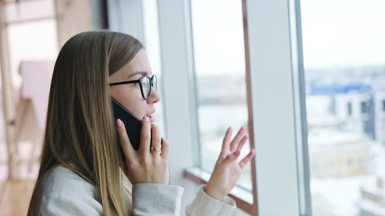 Blonde young woman talks to someone on the phone and waves her hand. Portrait of a happy confident lady standing in front of the window.