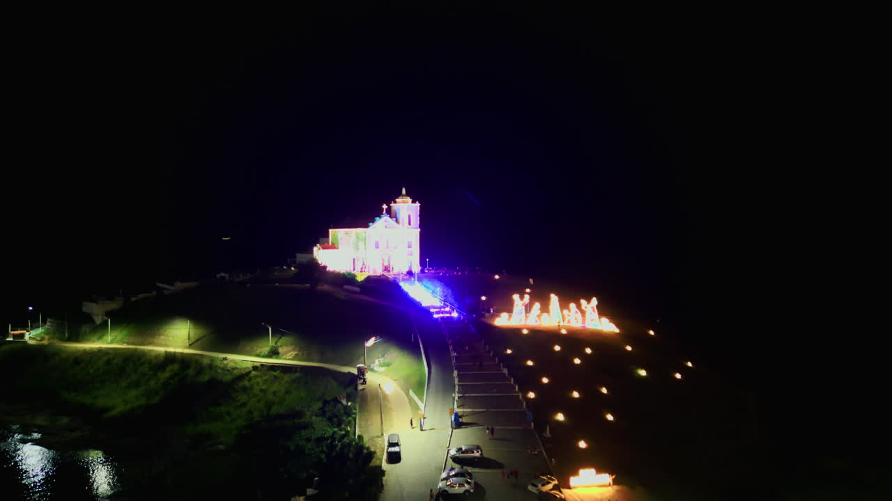Drone che vola in avanti sopra case residenziali verso la Chiesa di Nossa Senhora de Nazareth, illuminata a giorno sulla collina. Saquarema, Rio de Janeiro, Brasile. Luci di Natale. Video 4K