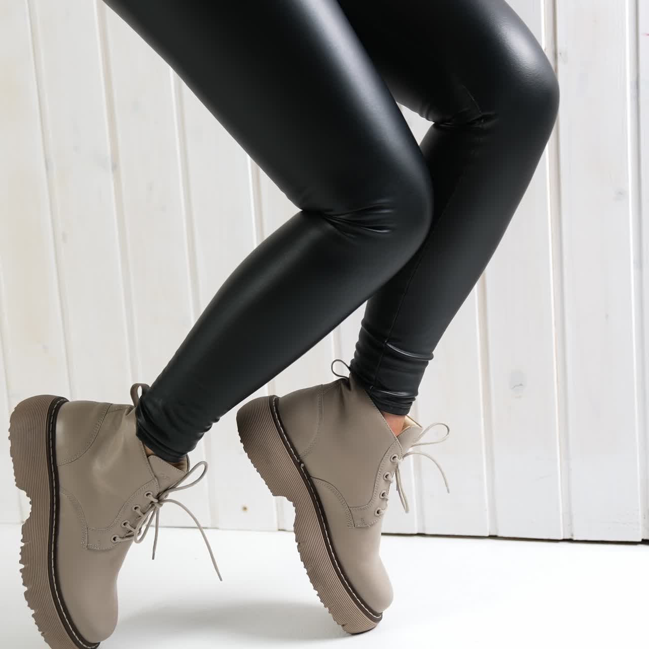 Demonstration of low beige boots with laces. Model in black leather pants jumps up in fashionable footwear. White wall backdrop