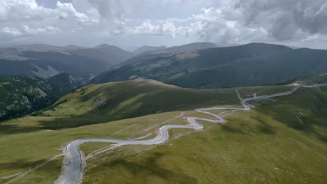 Breathtaking-view-of-the-winding-roads-of-Transalpina-in-Romania,-snaking-through-lush-green-meadows-against-a-backdrop-of-dramatic-mountain-peaks-and-moody-clouds