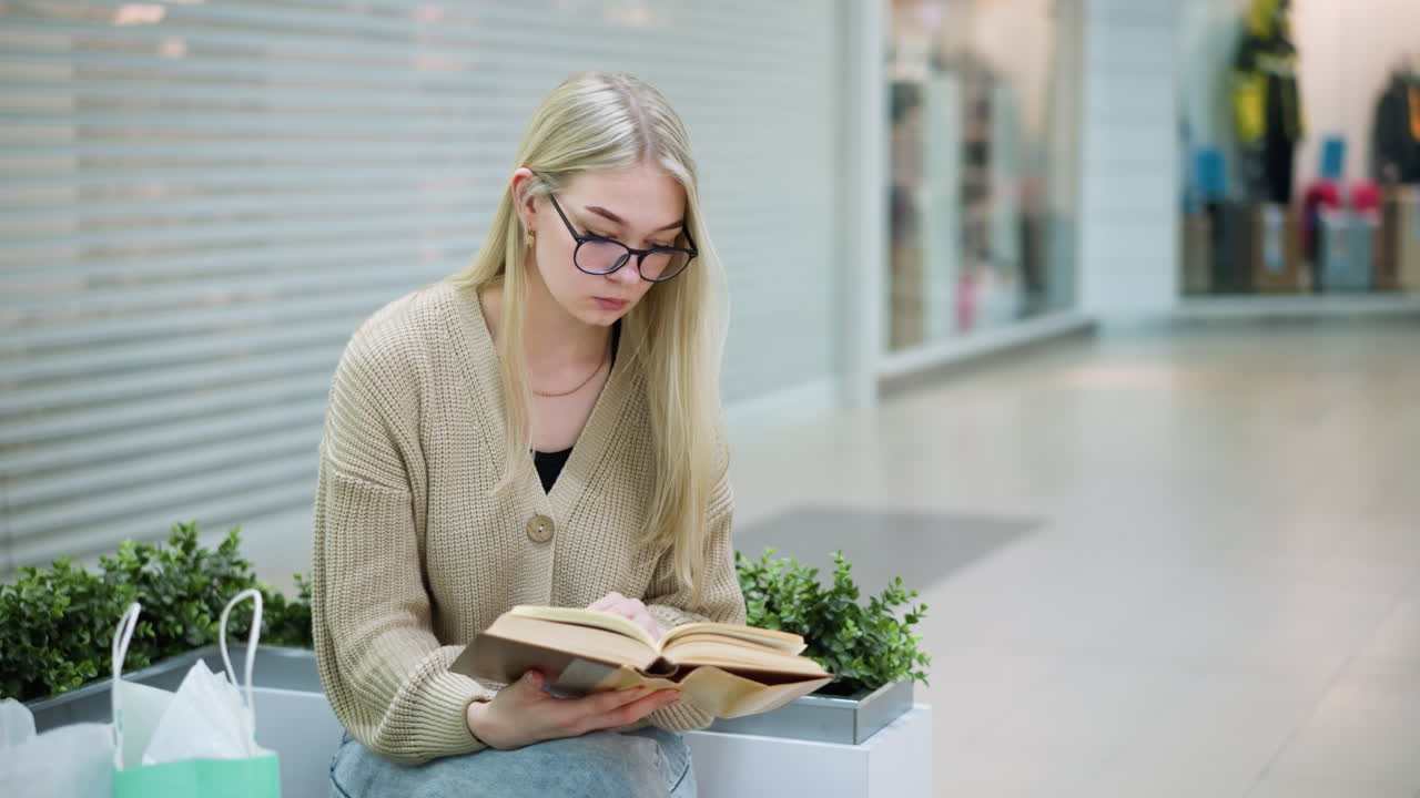 joven chica tranquila sentada en un centro comercial brillante con las piernas cruzadas, leyendo un libro, bolsas de compras cerca, tienda visible en el fondo, relajada, ambiente elegante perfecto para el ocio y las compras