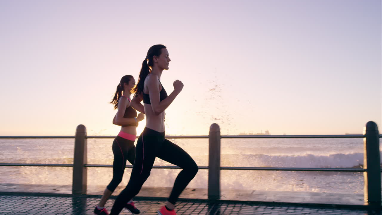 Due donne atletiche che corrono all'aperto al rallentatore sul lungomare al tramonto vicino all'oceano, godendosi la corsa serale.