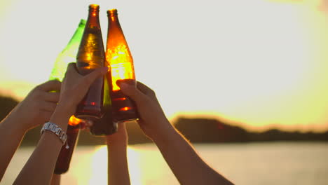 teenagers are dancing with hands up and clink beer from glass colorful bottles. this is perfect party at sunset on the lake coast.