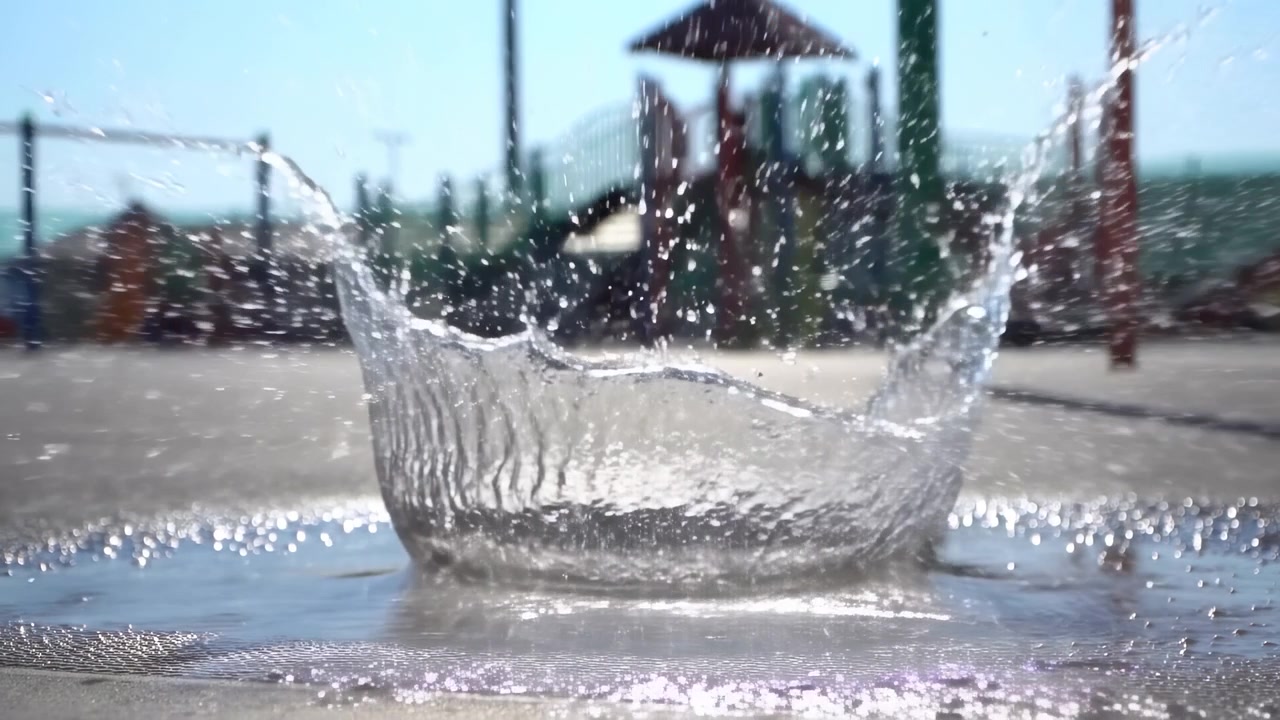 Bambini schizzano gioiosamente un pallone da calcio in una pozzanghera d'acqua su un parco giochi attrezzato con scivoli e altalene, catturato da una prospettiva all'altezza degli occhi, mostrando una scena dinamica e giocosa.