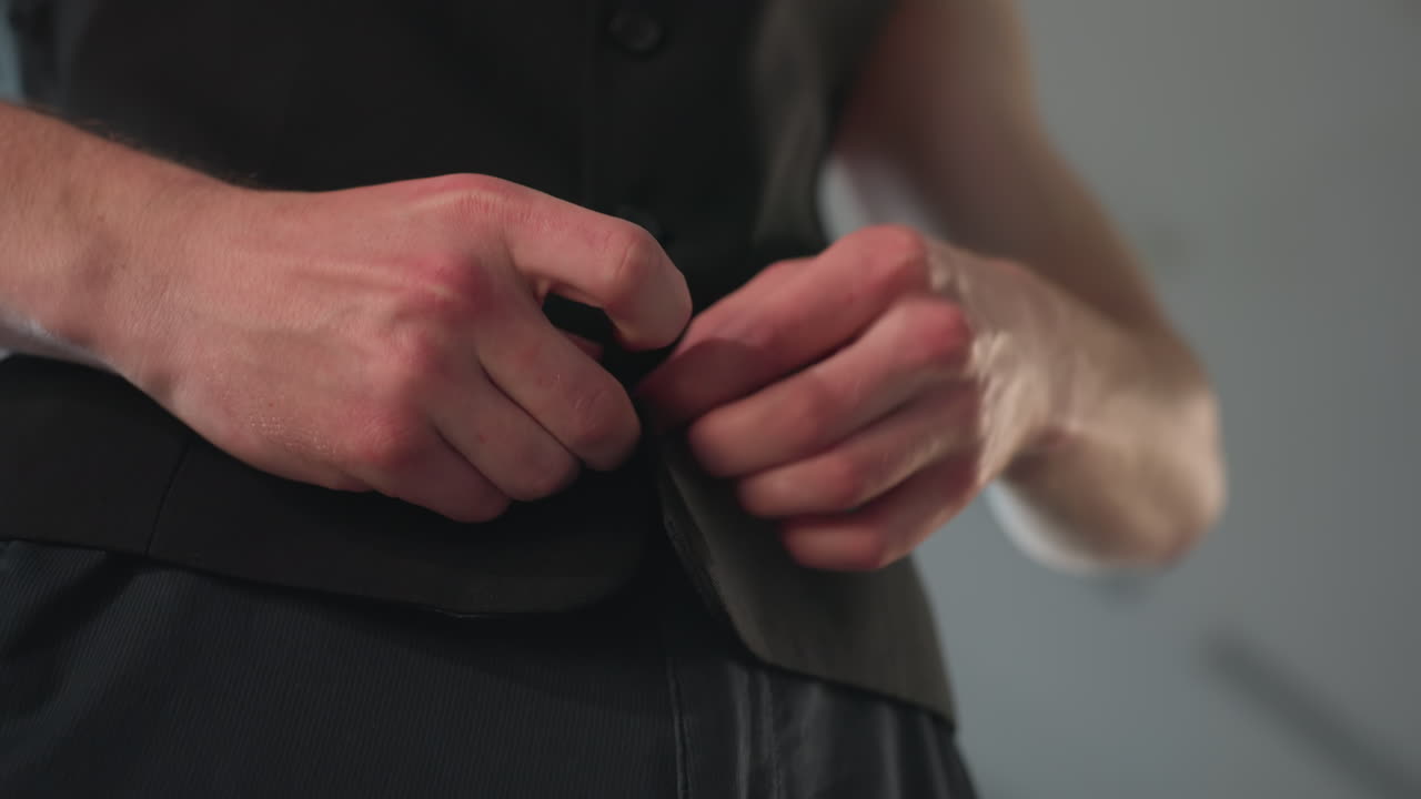 close up of man buttoning black jacket against blurred window light in modern loft interior shot, detail focused on hands fastening buttons for stylish formal attire preparation elegant