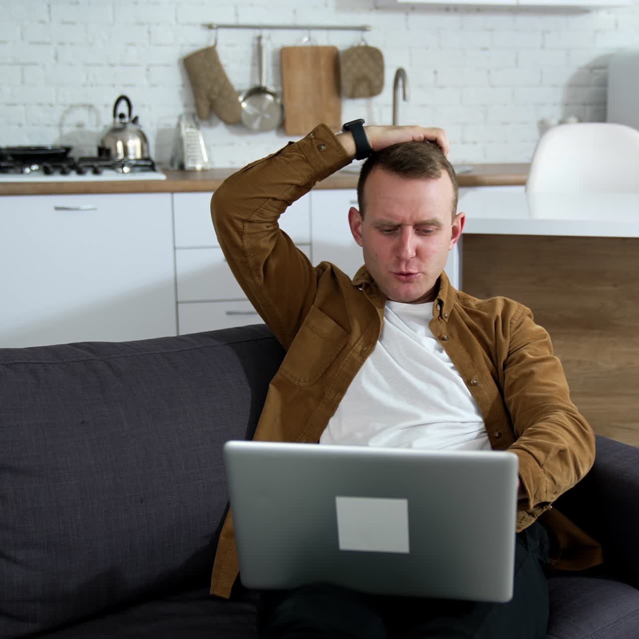 Young man playing computer games at home. Nervous guy laying on a couch with a laptop on kitchen background