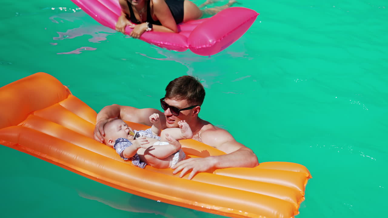 Daddy is holding a mattress with his baby on standing in the pool. Mother is swimming on the pink mattress at backdrop.