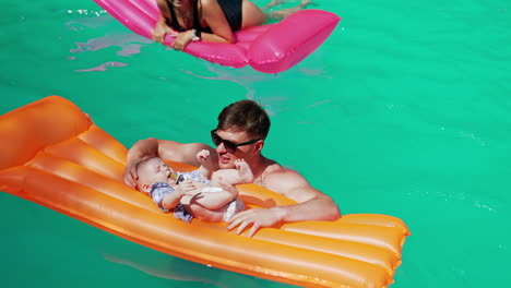 daddy is holding a mattress with his baby on standing in the pool. mother is swimming on the pink mattress at backdrop.