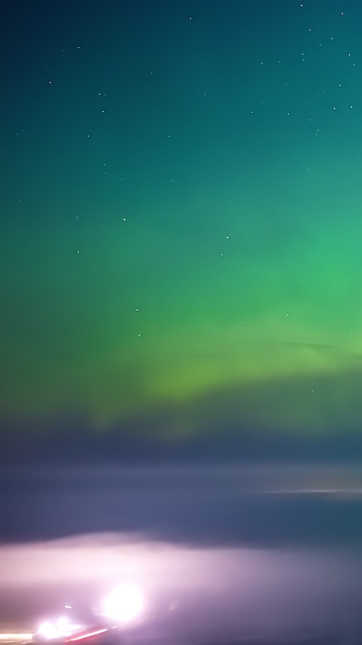 Splendido timelapse aereo verticale di intense luci verdi del nord, aurora boreale, che ondeggiano nel cielo notturno stellato su un paesaggio nebbioso con luci lontane nell'inverno in Lettonia